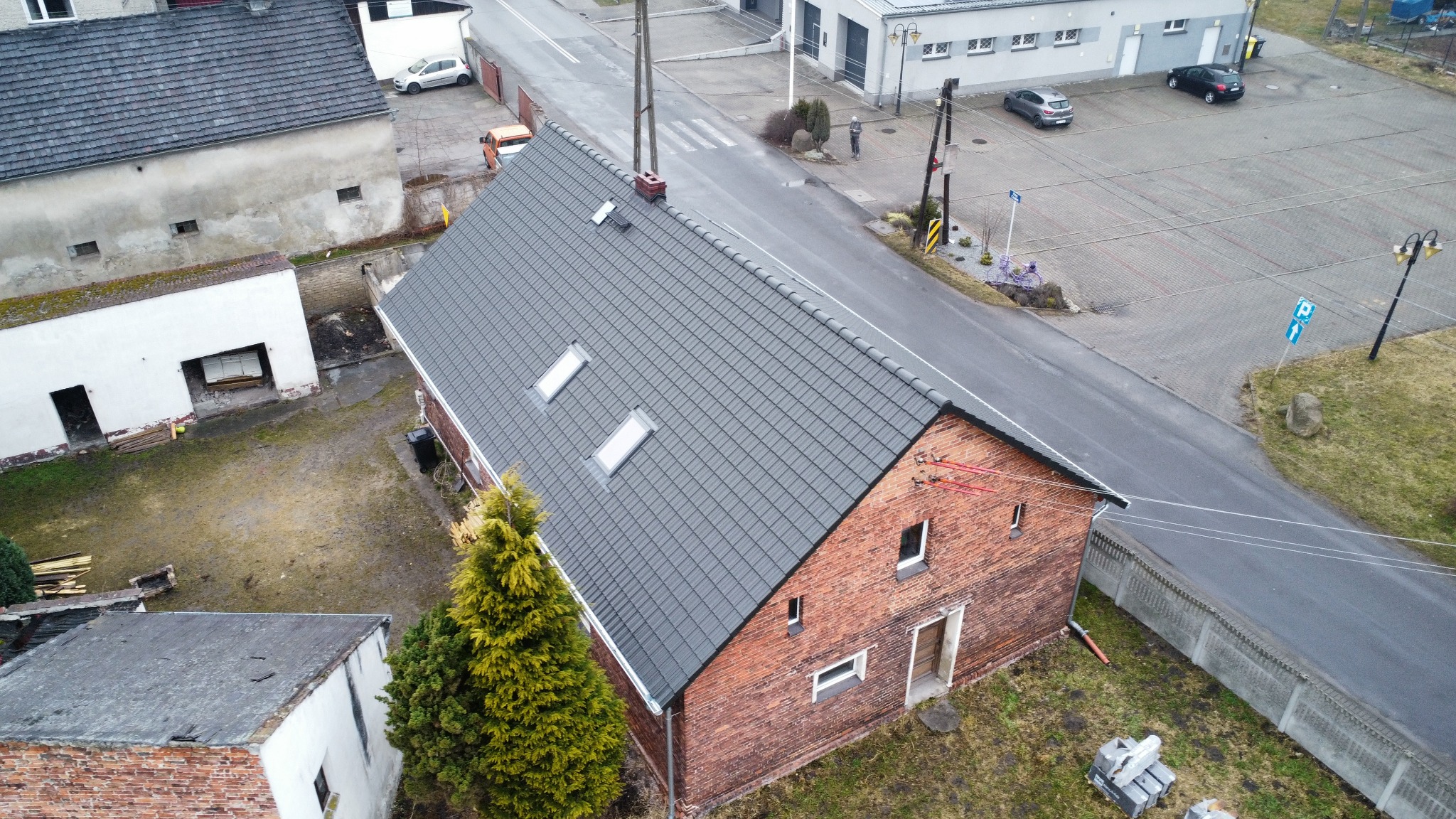 Ceglany dom z nowym, szarym dachem z dwoma oknami dachowymi, widziany z góry. W tle ulica i inne budynki. Dachówka ułożona równo, widoczne detale wykończenia.