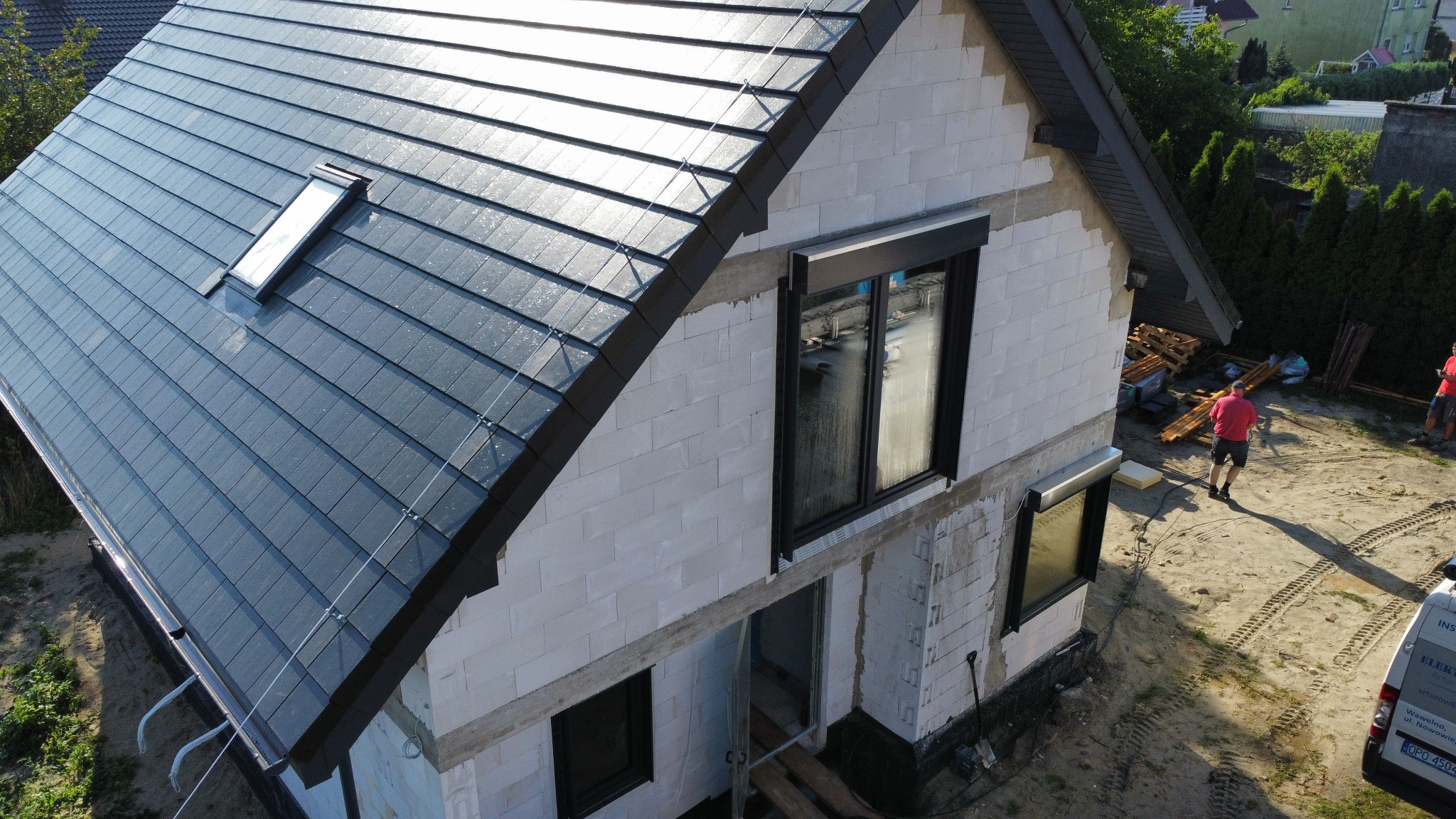 Nowoczesny dom w trakcie budowy z ciemnoszarą dachówką i oknem dachowym. Widoczne białe ściany z betonu komórkowego i czarne ramy okienne. Ujęcie z góry.