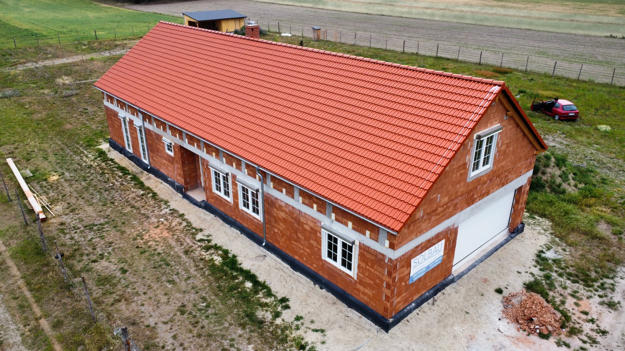 Budynek w trakcie budowy z czerwoną dachówką, widok z góry. Częściowo otynkowane ściany, okna wstawione, teren wokół budynku przygotowany. Widoczna tablica z nazwą firmy.