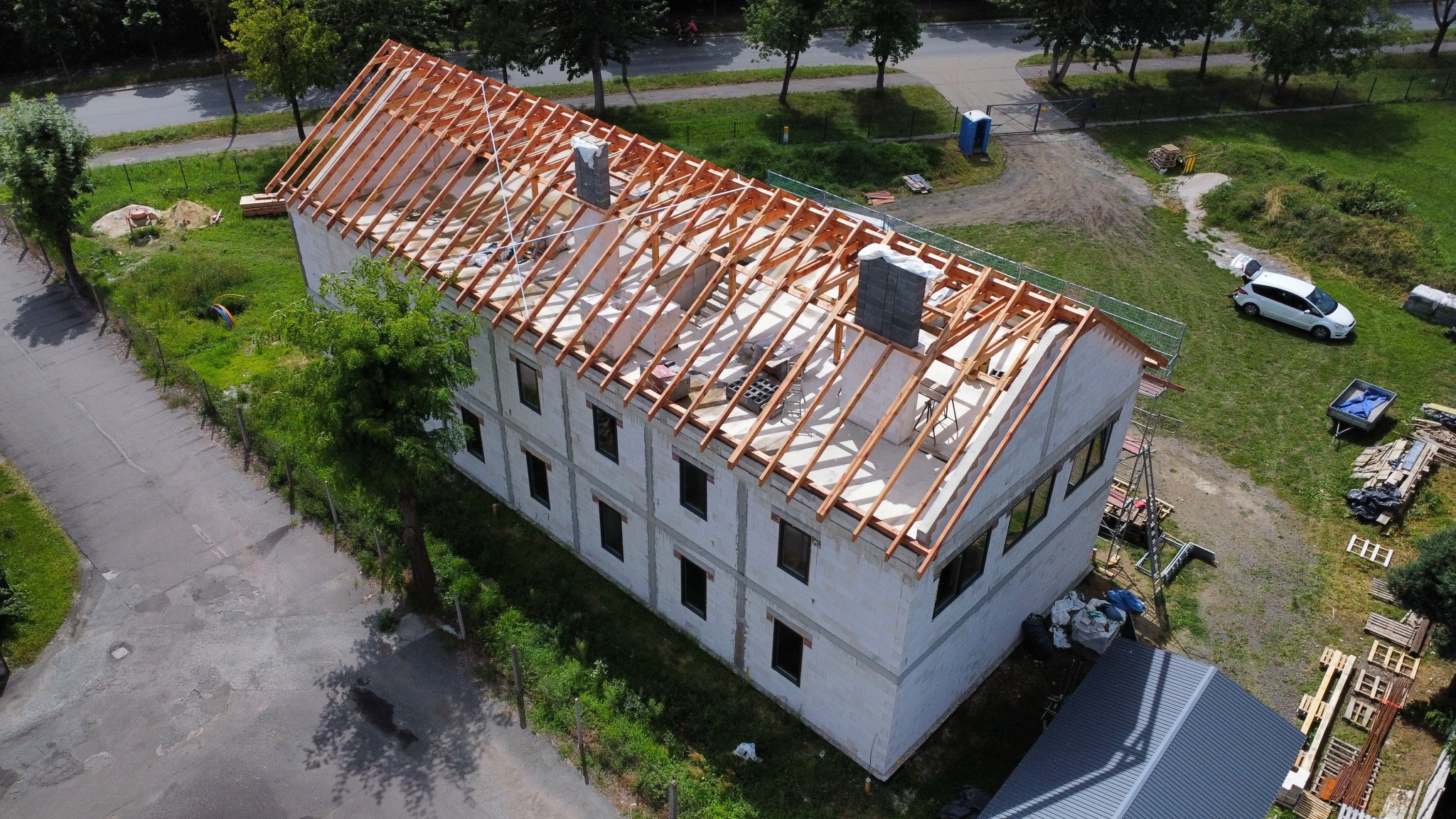 Budynek w trakcie budowy dachu, widok z góry. Elementy więźby dachowej z drewna, kominy z szarej cegły. Na trawniku zaparkowany biały samochód.