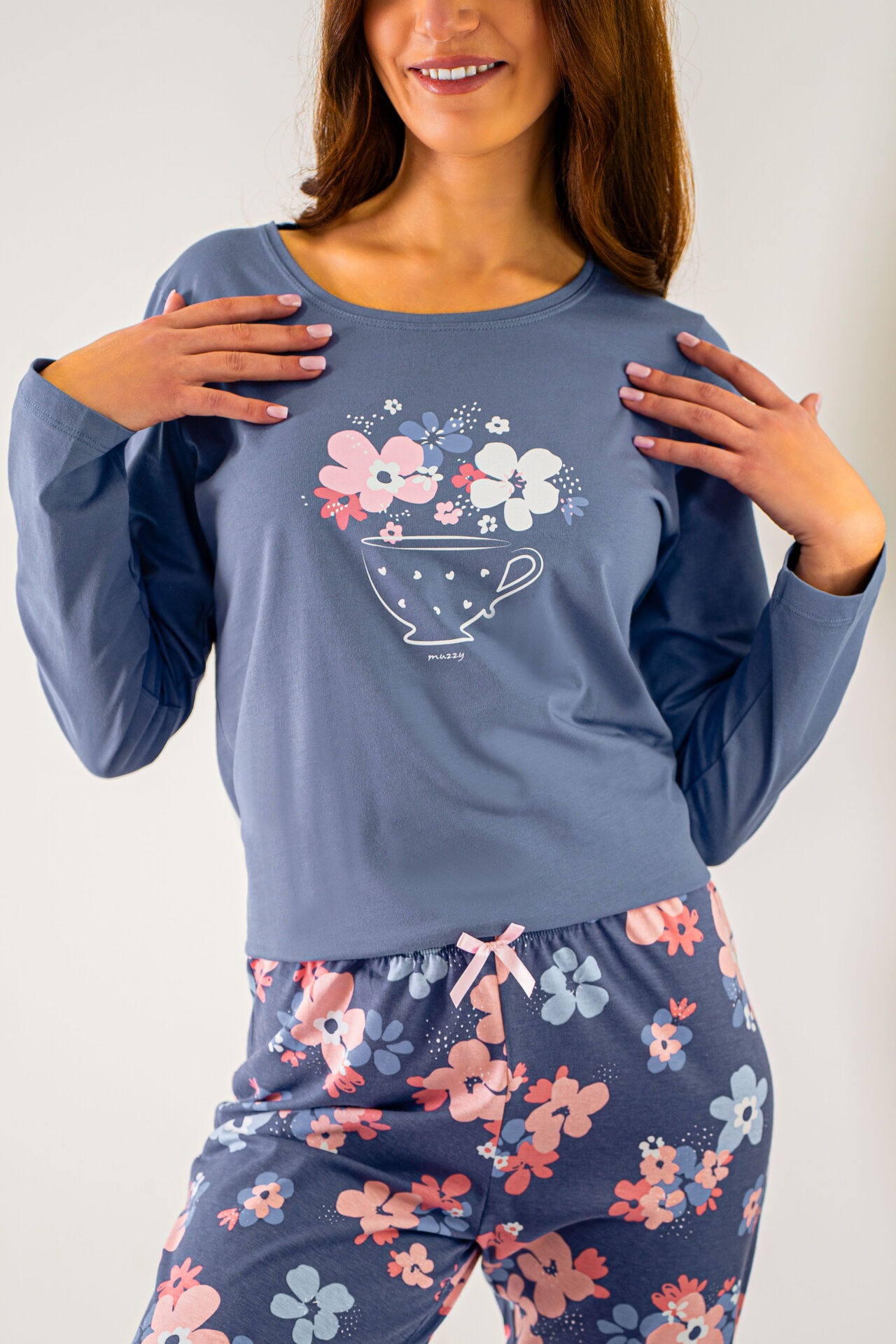 Kobieta w niebieskiej piżamie z długim rękawem i kwiatowym wzorem, prezentująca nadruk filiżanki z kwiatami. Różowa kokardka w pasie spodni.