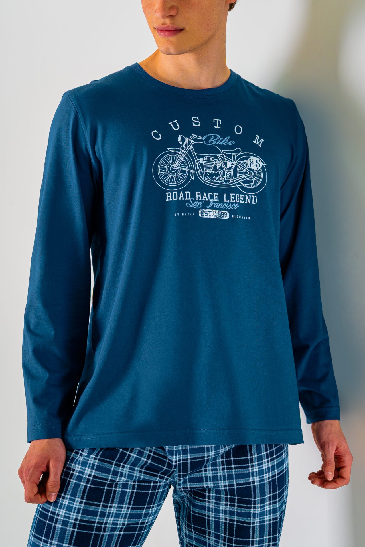 Mężczyzna w granatowej koszulce z długim rękawem i nadrukiem motocykla oraz w piżamie w kratę, stojący na tle białej ściany. Ujęcie od pasa w górę.