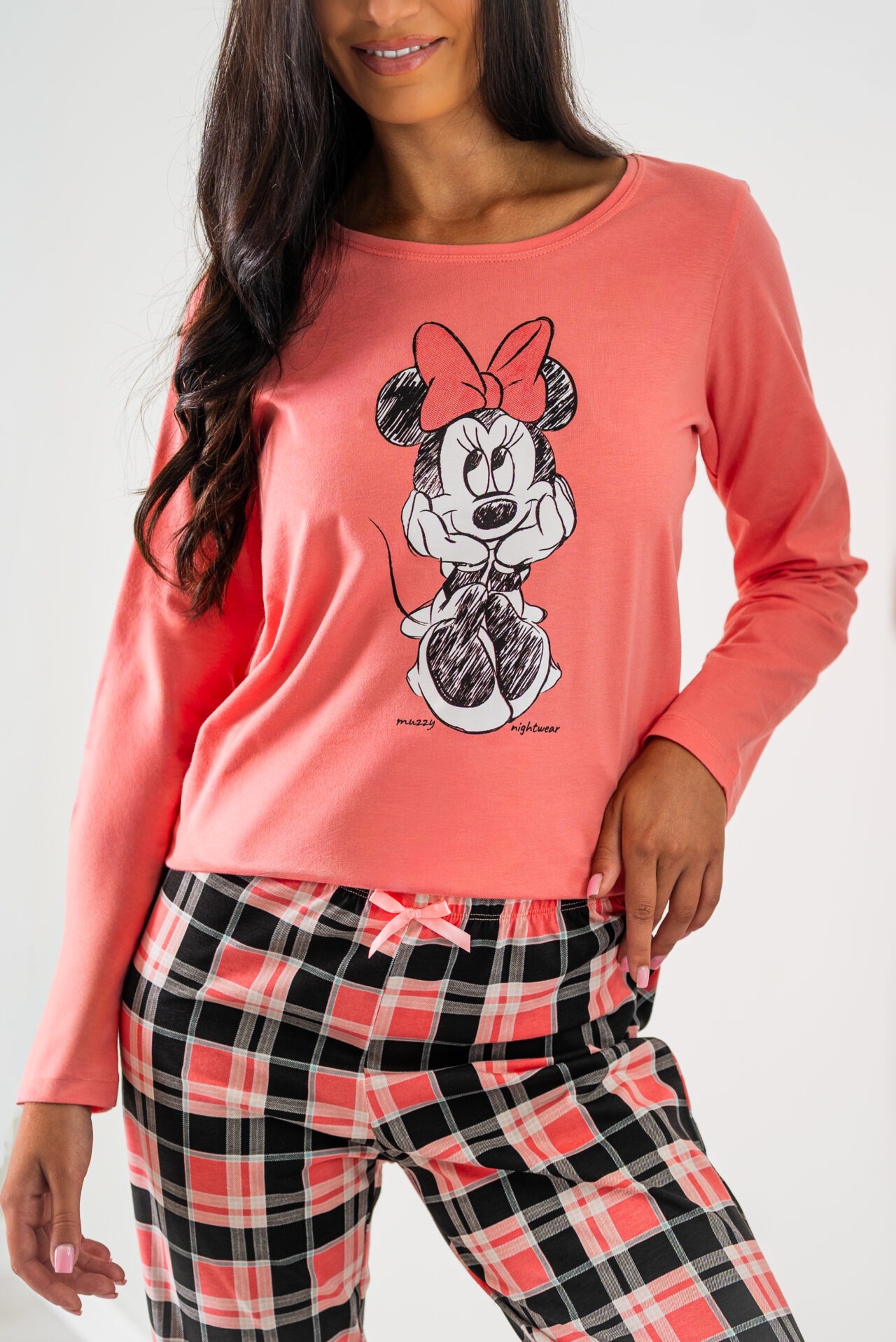 Kobieta w piżamie: koralowa bluzka z długim rękawem i nadrukiem Myszki Minnie, spodnie w kratę. Ujęcie od pasa w górę, na jasnym tle.