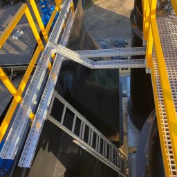VoltEco usługi elektryczne Patryk Kubulus - Widok z góry na konstrukcję z żółtymi barierkami i drabinką, z szarymi korytkami kablowymi, prowadzącymi do zbiornika. Elementy instalacji przemysłowej.