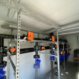 VoltEco usługi elektryczne Patryk Kubulus - Wnętrze pomieszczenia z rozbudowaną instalacją rur, zaworów (niebieskich i czarnych) i automatyki (pomarańczowej), wspartych na metalowych konstrukcjach. Widoczne odbicia na podłodze.