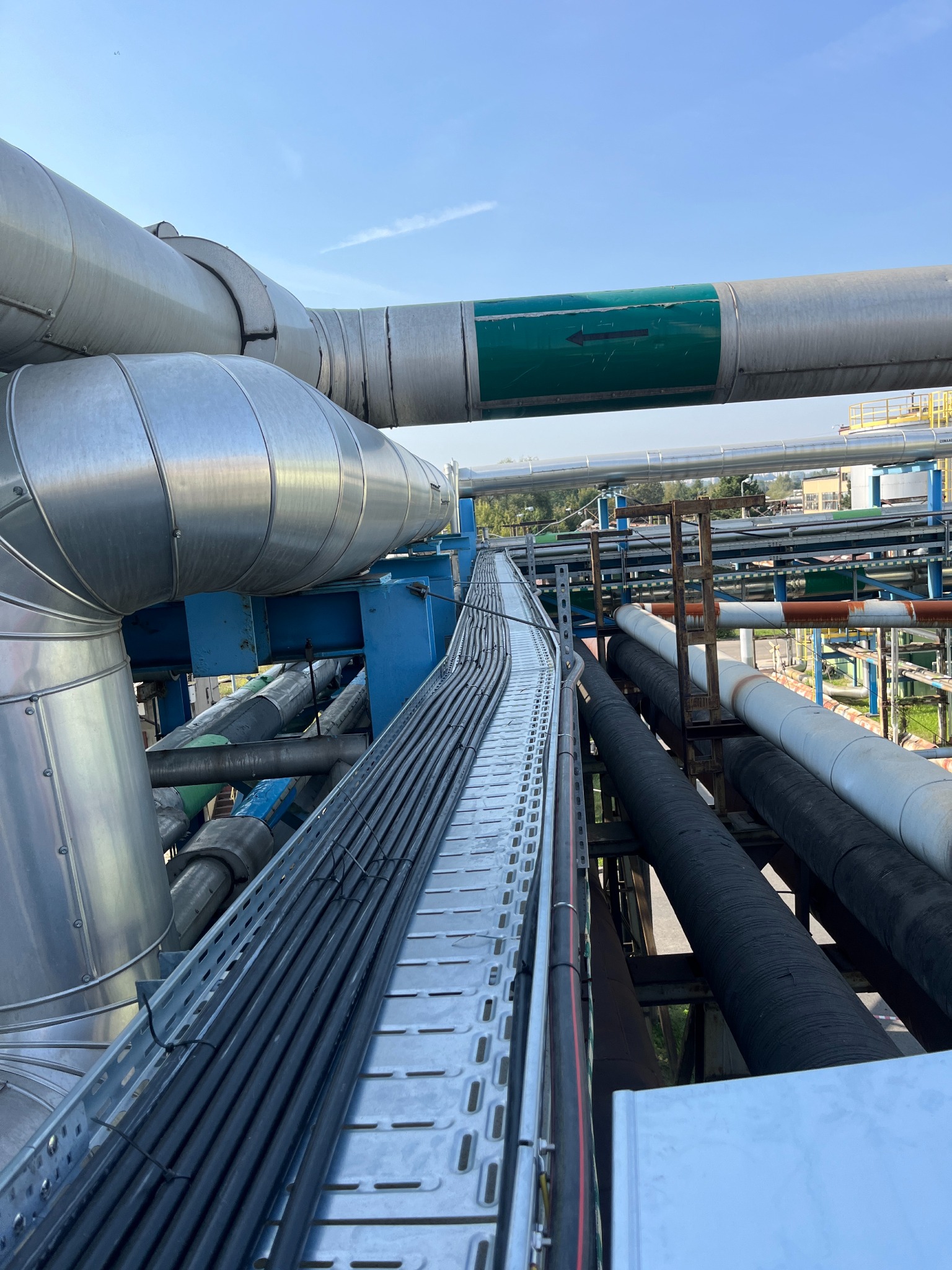 Metalowa korytka kablowe z przewodami, zamocowane na konstrukcji z rurami w tle. Widok z góry, oświetlenie dzienne, błękitne niebo w tle.