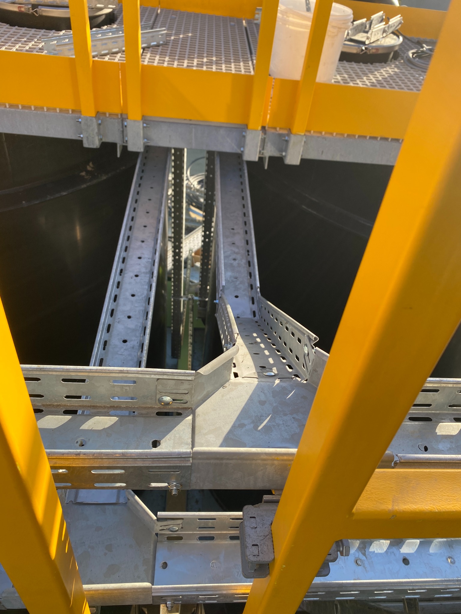 Metalowe korytka kablowe zamontowane na konstrukcji z żółtymi barierkami, widok z góry. Elementy instalacji elektrycznej w przestrzeni przemysłowej.