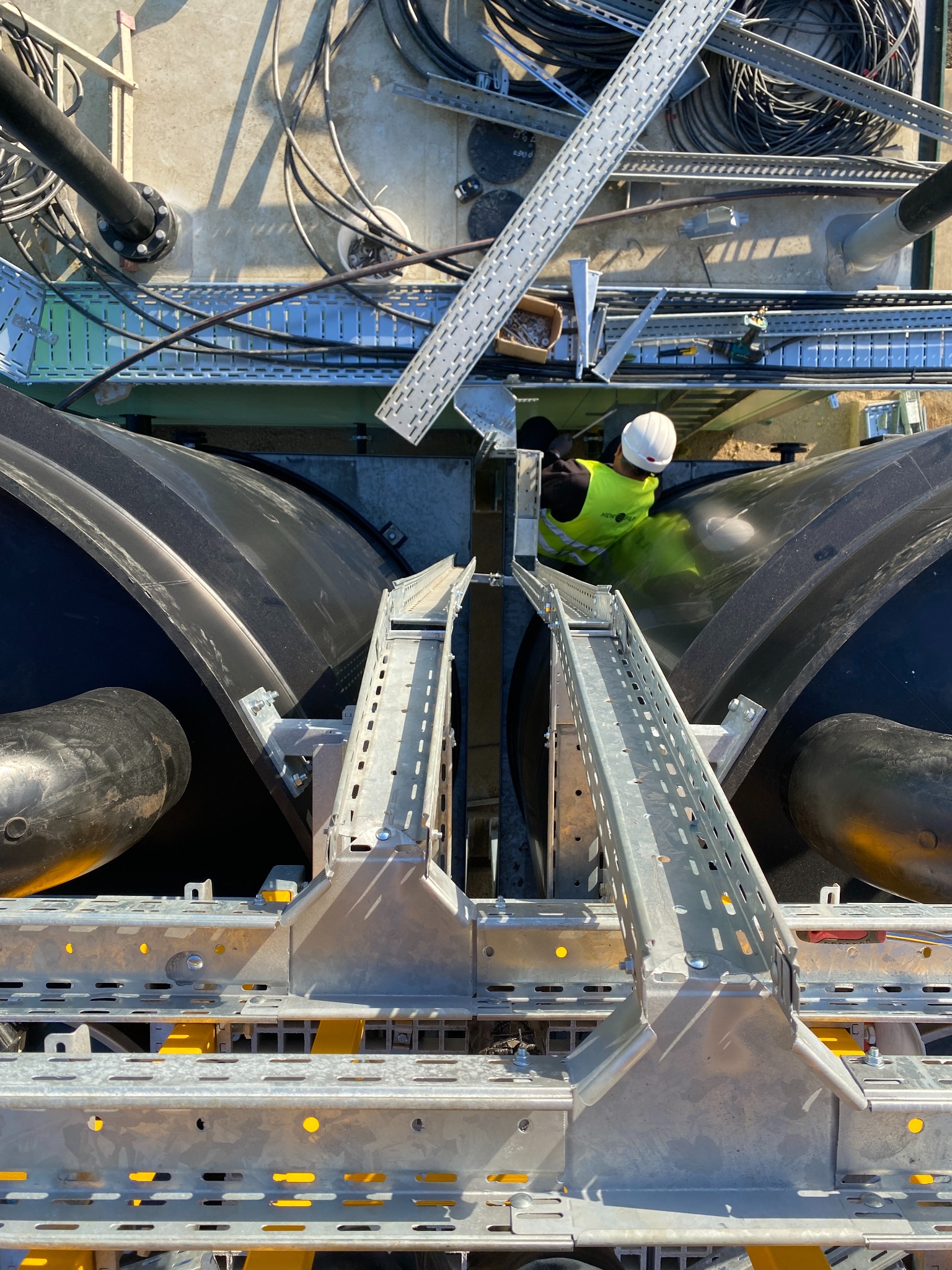 Praca na wysokości: instalacja korytek kablowych i rur na konstrukcji zbiornika. Pracownik w kasku i kamizelce odblaskowej. Widok z góry, ostre światło.