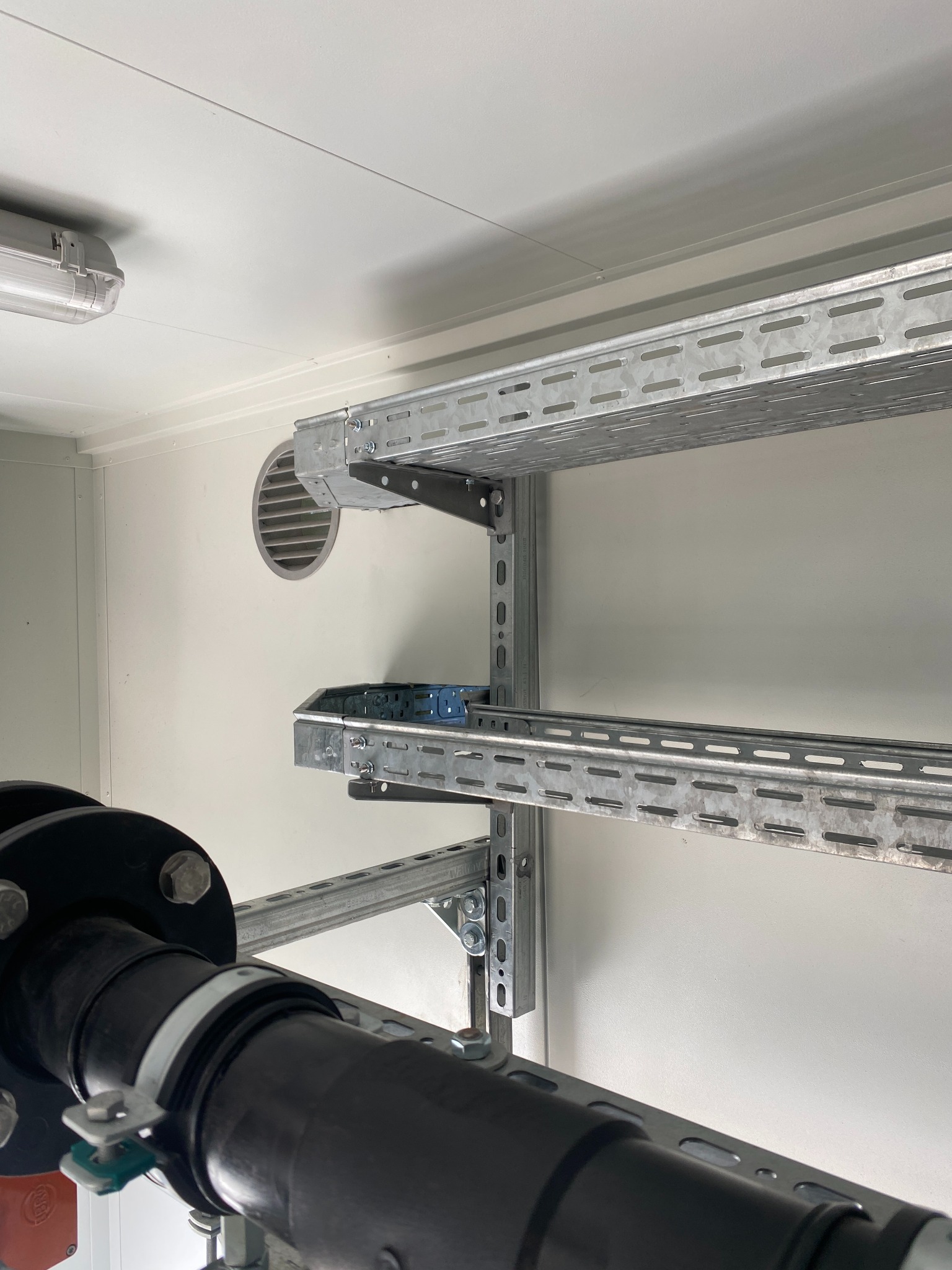 Metalowe korytka kablowe zamocowane do ściany, widoczny fragment rury z kołnierzem i wentylacja. Białe ściany i sufit, oświetlenie jarzeniowe.