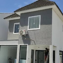 JS BRASIL - Elewacja domu po malowaniu w Poznaniu. Widoczny pracownik na drabinie, okna z białymi ramami, klimatyzator i basen. Szara fasada budynku.