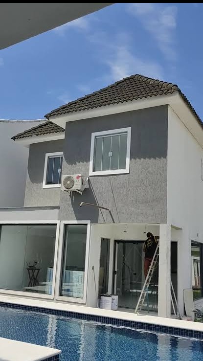 Elewacja domu po malowaniu w Poznaniu. Widoczny pracownik na drabinie, okna z białymi ramami, klimatyzator i basen. Szara fasada budynku.