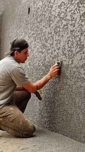 Nakładanie struktury na ścianę zewnętrzną przez mężczyznę w czapce, używającego pacy. Szara ściana z wyraźną fakturą, widoczne narzędzie i fragment sylwetki wykonawcy.
