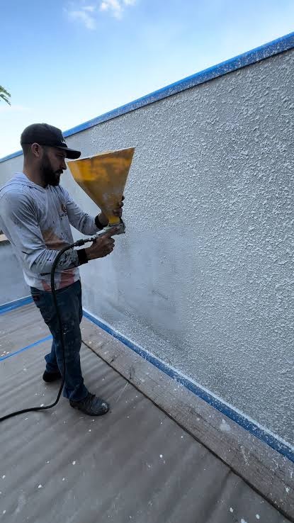 Malowanie natryskowe ściany zewnętrznej przez mężczyznę w czapce. Widoczny pistolet natryskowy i świeżo nałożona, chropowata faktura farby. Niebieskie zabezpieczenie krawędzi.