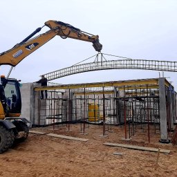 CONSTRUCTOR Zbudujmy - Żuraw CAT podnosi zbrojenie łukowe nad budową z bloczków betonowych. Widoczny pracownik na konstrukcji. Tło: pole i pochmurne niebo. Etap budowy domu.