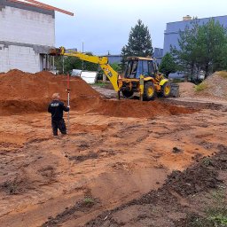 CONSTRUCTOR Zbudujmy - Żółta koparka JCB przygotowuje teren pod budowę domu. Geodeta z niwelatorem kontroluje poziom wykopu. W tle widać budynek w stanie surowym otwartym.