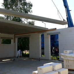 CONSTRUCTOR Zbudujmy - Budowa domu z betonu komórkowego: montaż prefabrykowanych stropów za pomocą dźwigu. Widoczne ściany, otwory okienne i paleta z bloczkami.