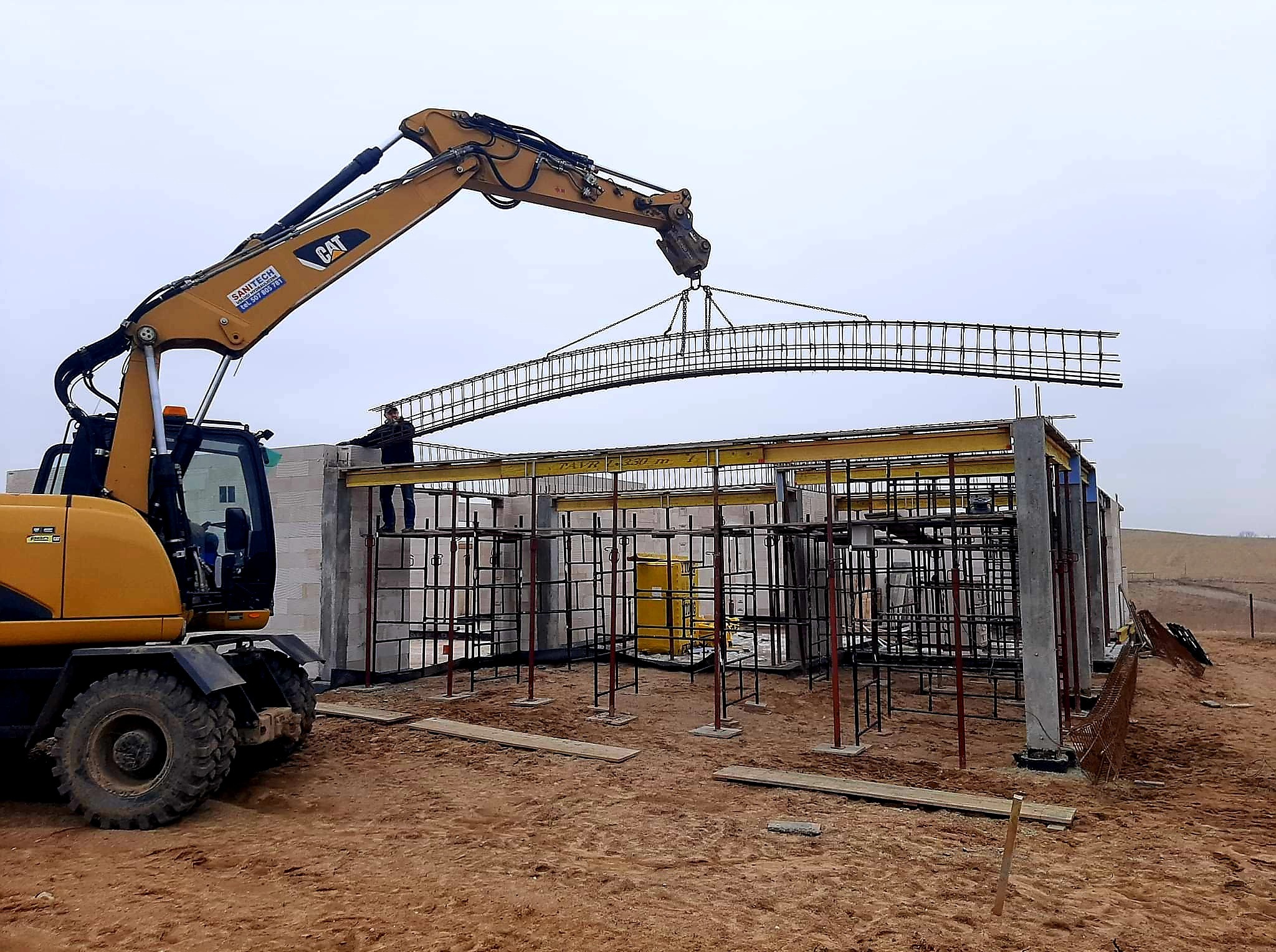 Żuraw CAT podnosi zbrojenie łukowe nad budową z bloczków betonowych. Widoczny pracownik na konstrukcji. Tło: pole i pochmurne niebo. Etap budowy domu.