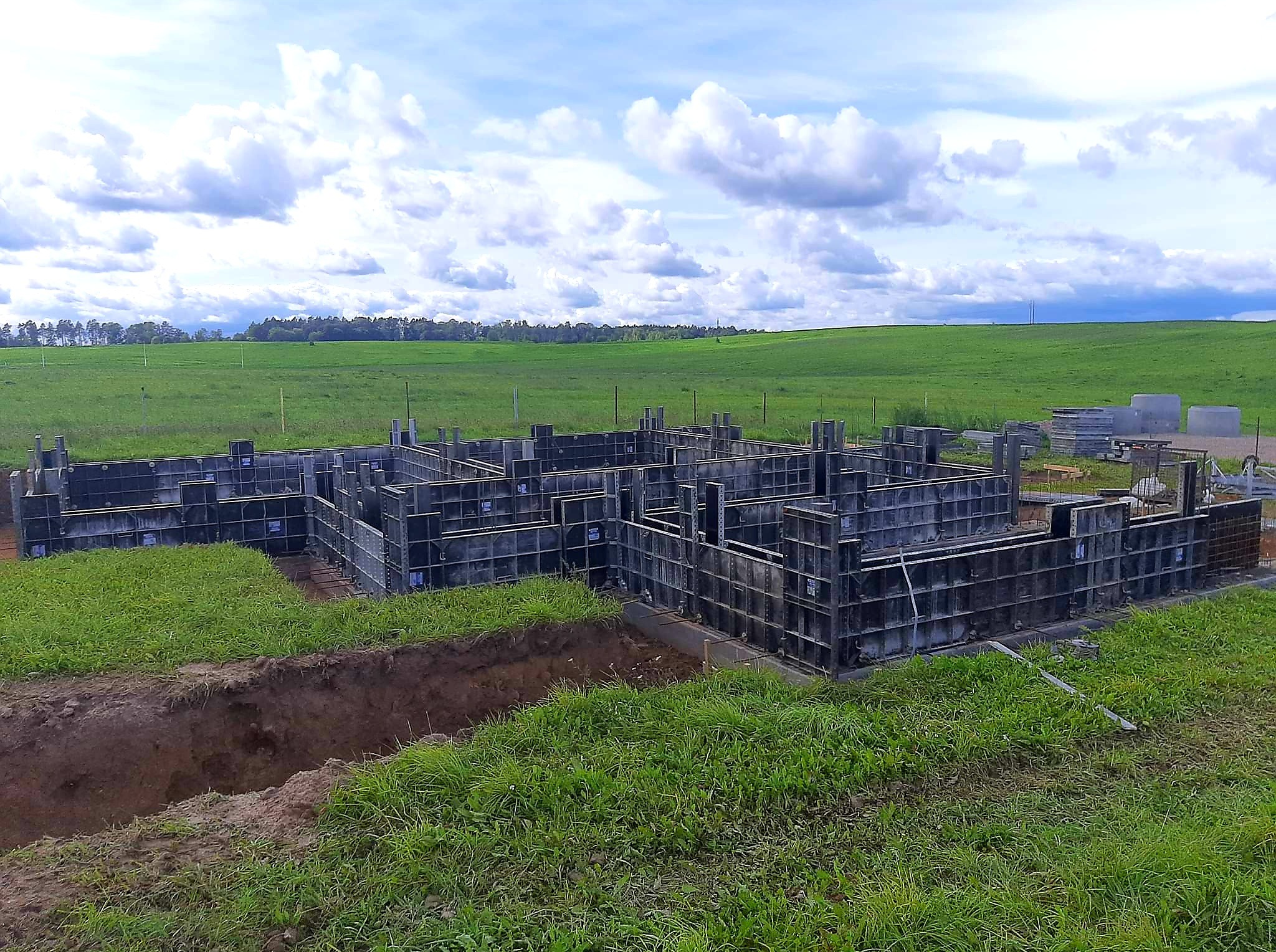Szare szalunki fundamentowe na zielonej trawie pod błękitnym niebem z chmurami. Widoczny wykop pod fundament. Krajobraz wiejski w tle.