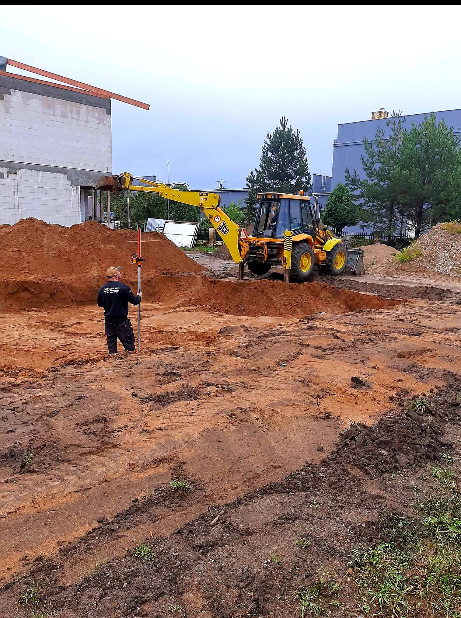 Żółta koparka JCB przygotowuje teren pod budowę domu. Geodeta z niwelatorem kontroluje poziom wykopu. W tle widać budynek w stanie surowym otwartym.