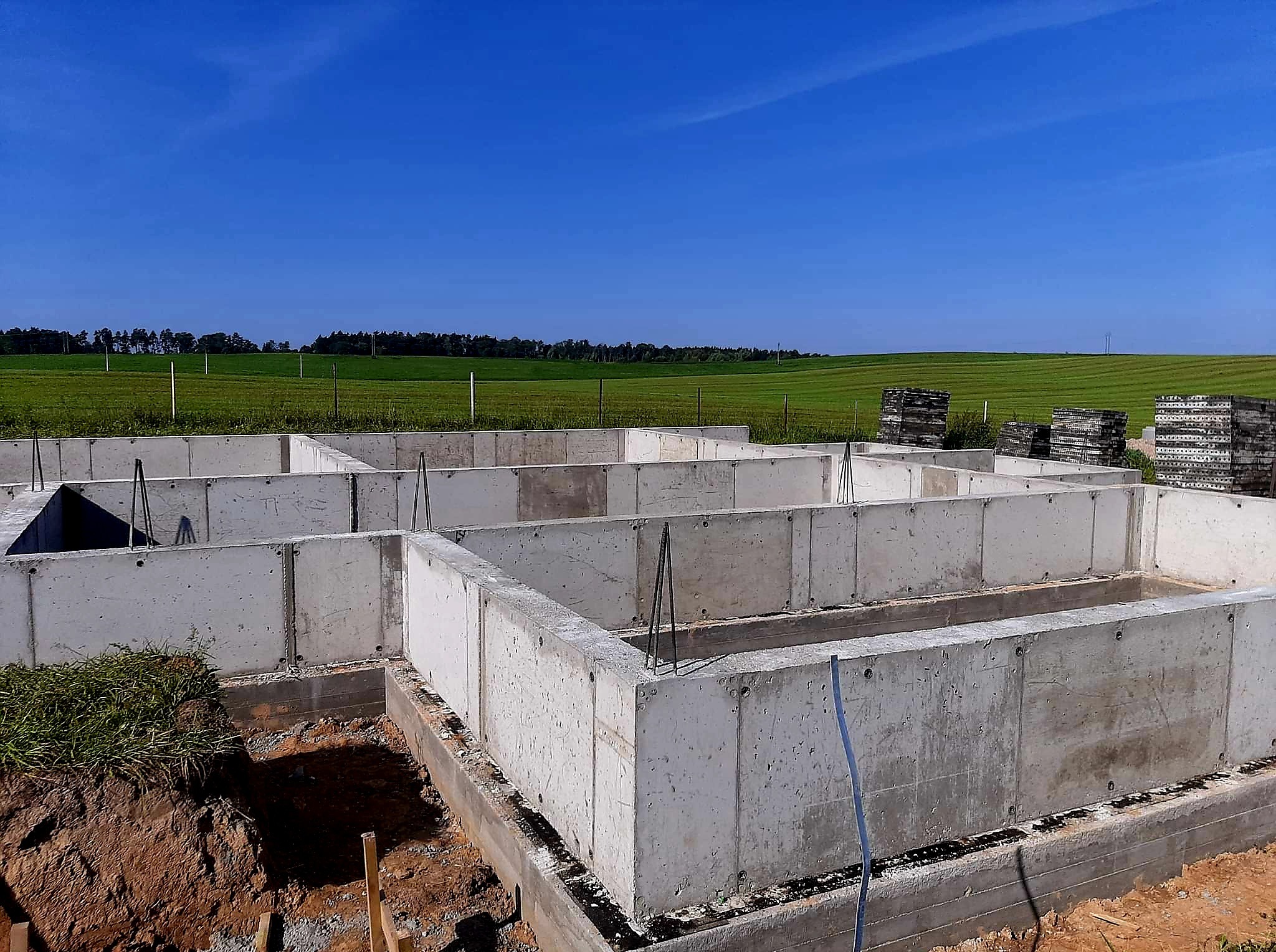 Betonowe fundamenty domu na tle zielonego pola i błękitnego nieba. Widoczne zbrojenia i elementy konstrukcyjne. Fundamenty wylane, gotowe do dalszych prac.