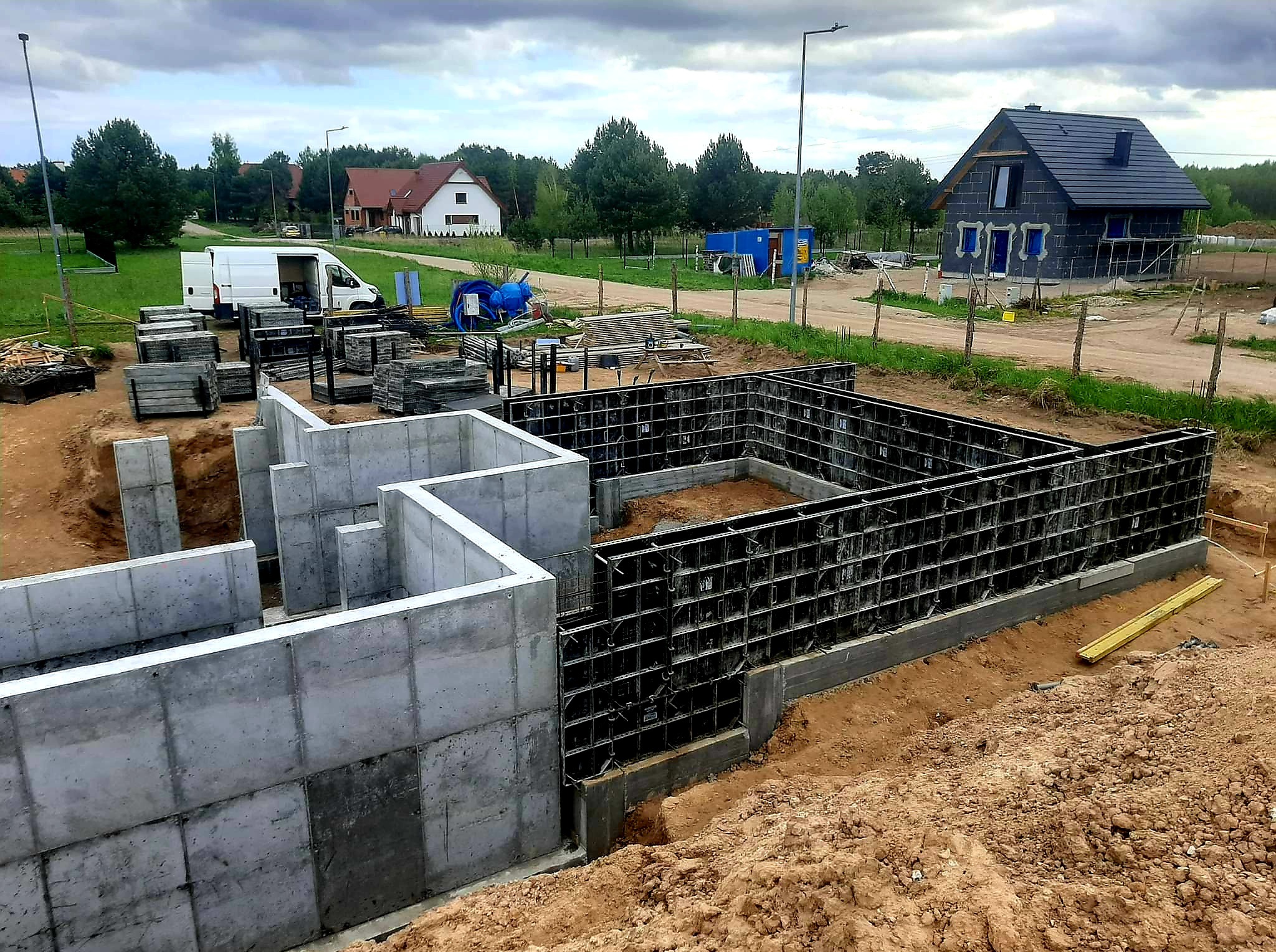 Fundamenty domu w trakcie budowy: betonowe ściany szalunkowe, widoczny teren budowy, częściowo wybudowany dom w tle, pochmurne niebo.