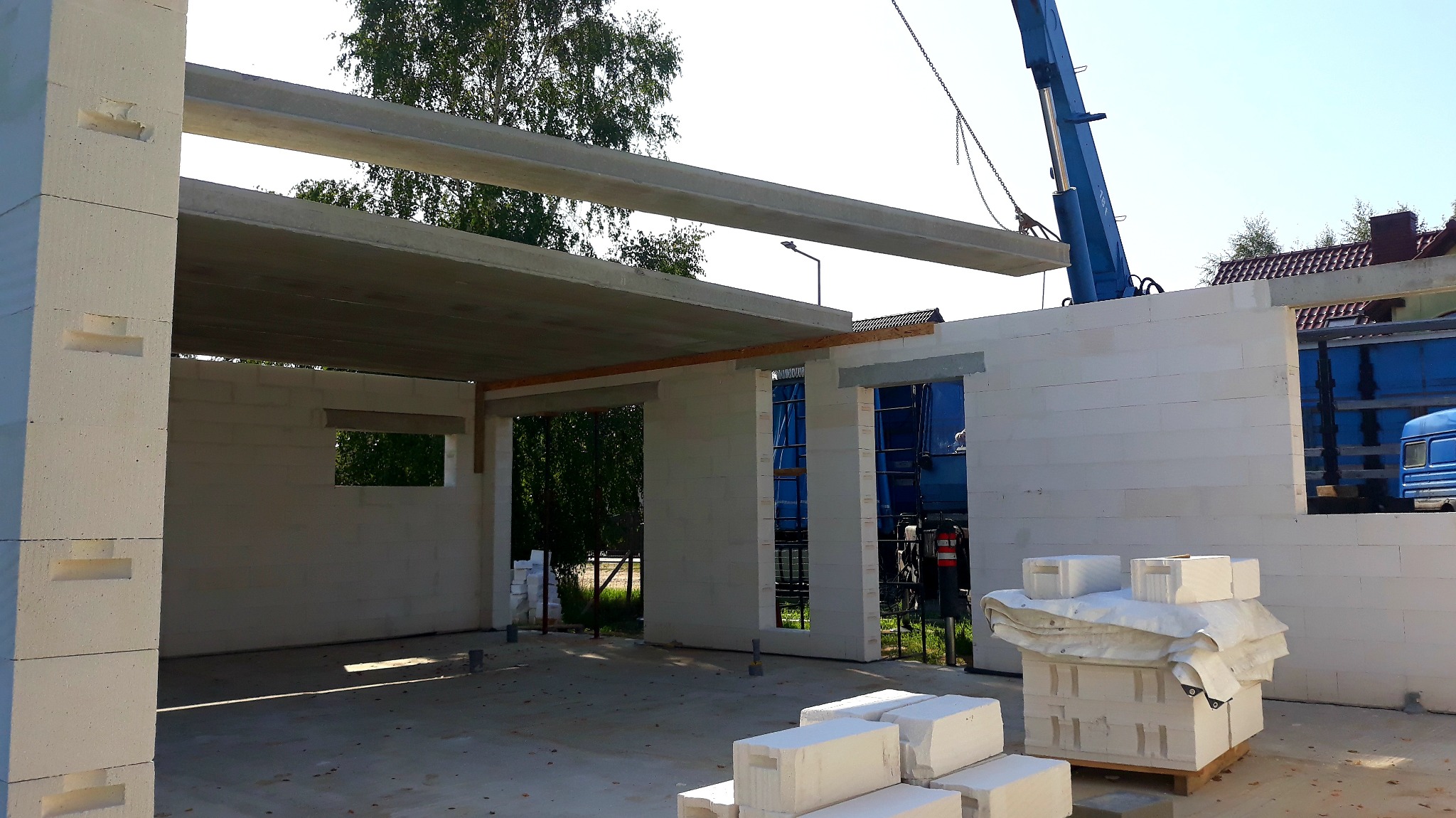 Budowa domu z betonu komórkowego: montaż prefabrykowanych stropów za pomocą dźwigu. Widoczne ściany, otwory okienne i paleta z bloczkami.