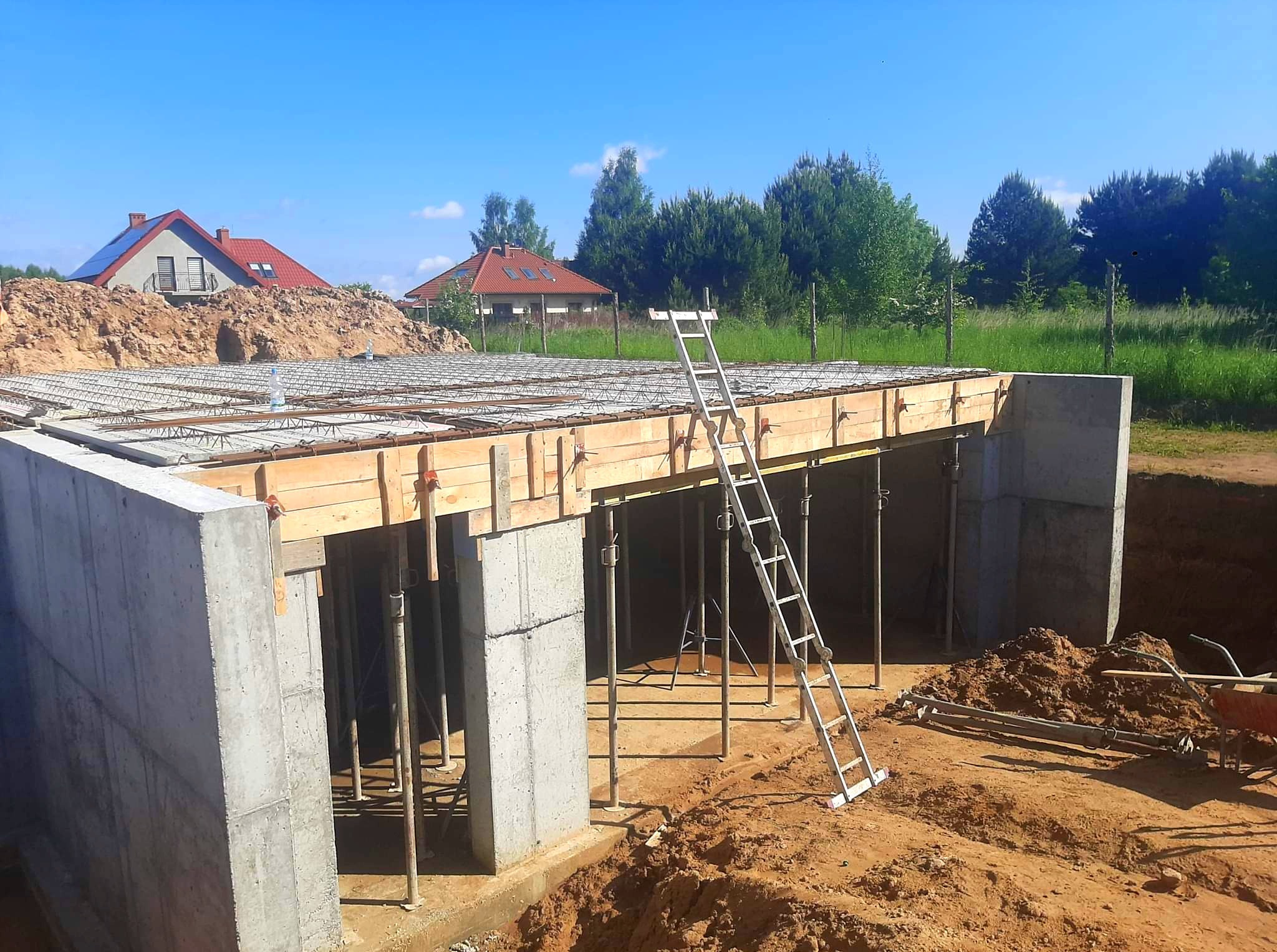 Zbrojona płyta fundamentowa z betonowymi ścianami, drewnianym szalunkiem i metalową drabiną na tle zieleni i błękitnego nieba. Widoczne domy w oddali.