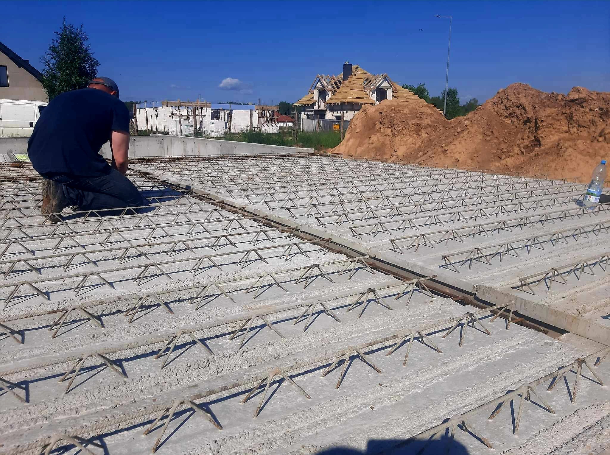 Zbrojenie płyty fundamentowej pod budowę domu jednorodzinnego. Widoczny pracownik w trakcie montażu, w tle budynek w stanie surowym otwartym i hałda ziemi.