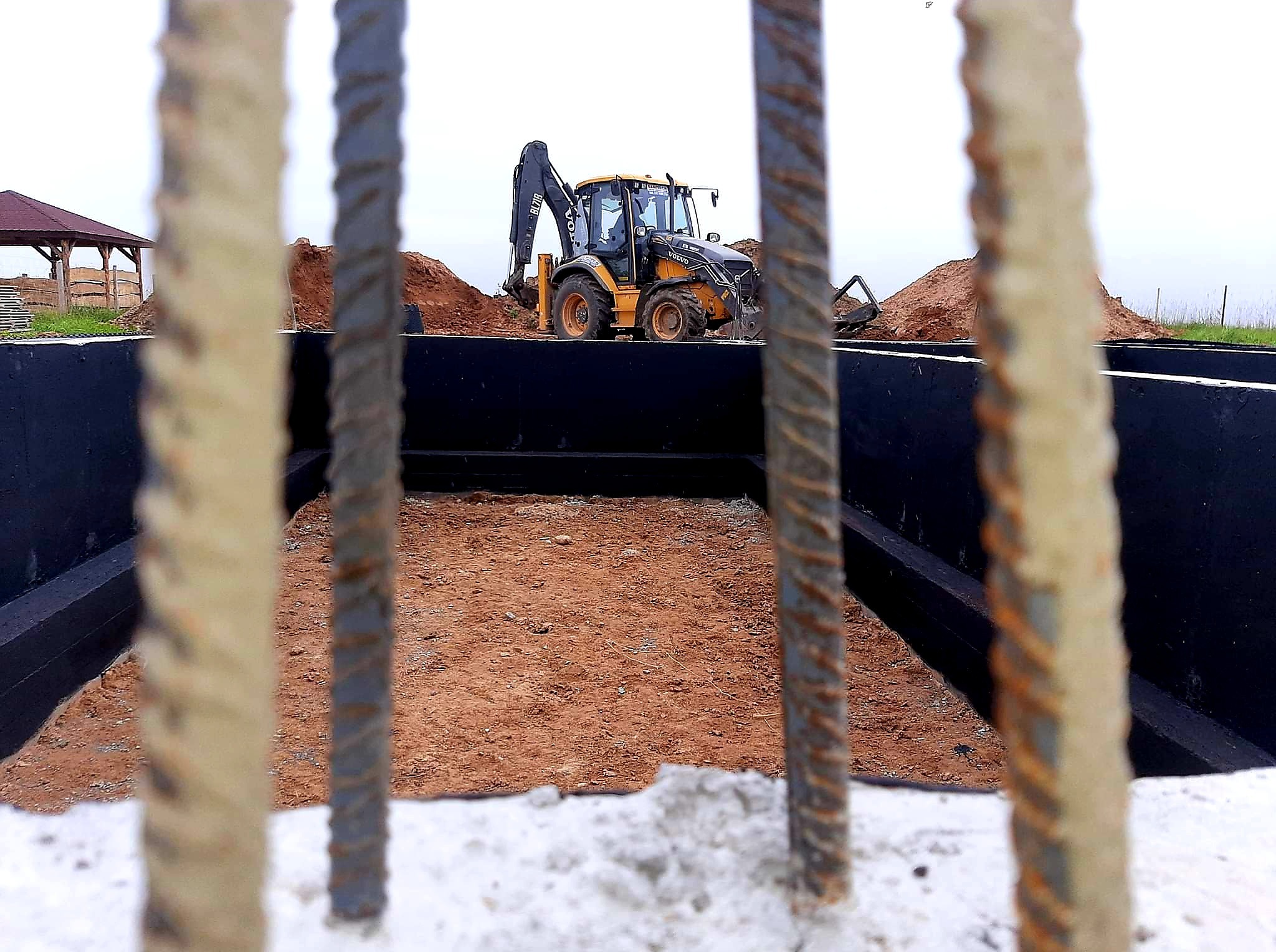 Wykop pod fundamenty z widocznym dnem wyłożonym piaskiem, zabezpieczonymi ścianami i koparką w tle, widziany przez pręty zbrojeniowe.