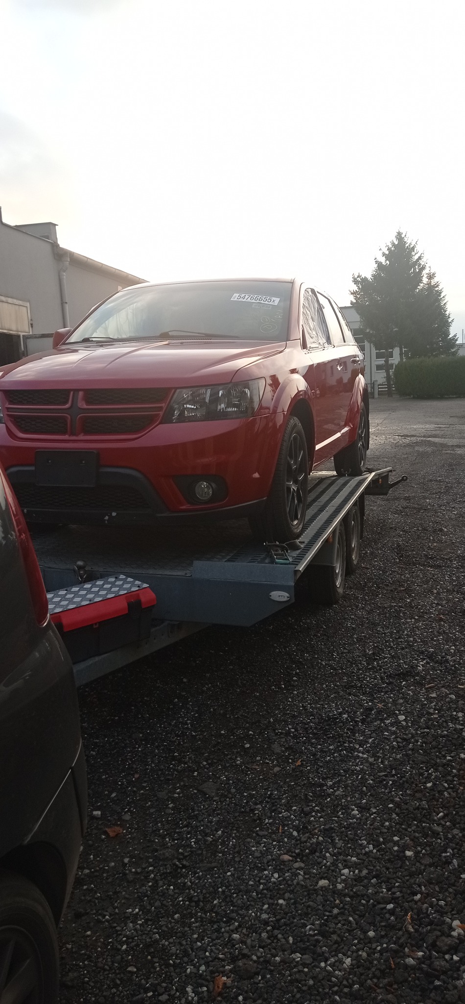 Czerwony Dodge na szarej lawecie, widok z boku, tabliczka z numerem na przedniej szybie, szare niebo w tle, częściowo widoczny inny pojazd.