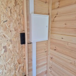 ELEKTRYK PRO JAKUB PAJĄK - Wnętrze drewnianego domu z zamontowaną białą skrzynką elektryczną i czarnymi gniazdkami. Widoczne płyty OSB i boazeria. Ujęcie z perspektywy narożnika.