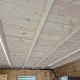ELEKTRYK PRO JAKUB PAJĄK - Drewniany sufit z zamontowanym oświetleniem LED wzdłuż belek. Widoczne kable i fragment ściany z płyty OSB. Ujęcie z dołu, szeroki kąt.