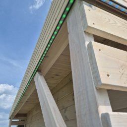 ELEKTRYK PRO JAKUB PAJĄK - Drewniana konstrukcja z oświetleniem LED w kolorze zielonym i niebieskim, zamontowanym pod dachem, na tle błękitnego nieba z chmurami. Ujęcie z dołu.