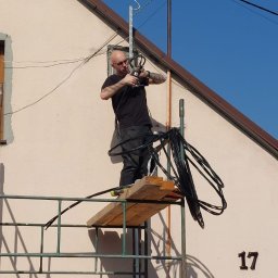 ELEKTRYK PRO JAKUB PAJĄK - Elektryk na rusztowaniu montuje kable na elewacji budynku z numerem 17. Widoczne narzędzia i instalacja antenowa. Słoneczny dzień, błękitne niebo.