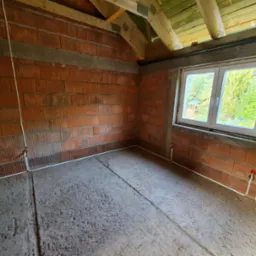 Surowy stan poddasza z instalacją elektryczną w peszlach przy ścianach z cegły, widoczne okno i drewniana konstrukcja dachu. Szara wylewka na podłodze.