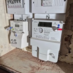 ELEKTRYK PRO JAKUB PAJĄK - Dwa liczniki elektryczne (GAMA 100 i OTUS 3) zamontowane obok skrzynki z bezpiecznikami wewnątrz budynku. Widoczne oznaczenia i numery seryjne urządzeń.