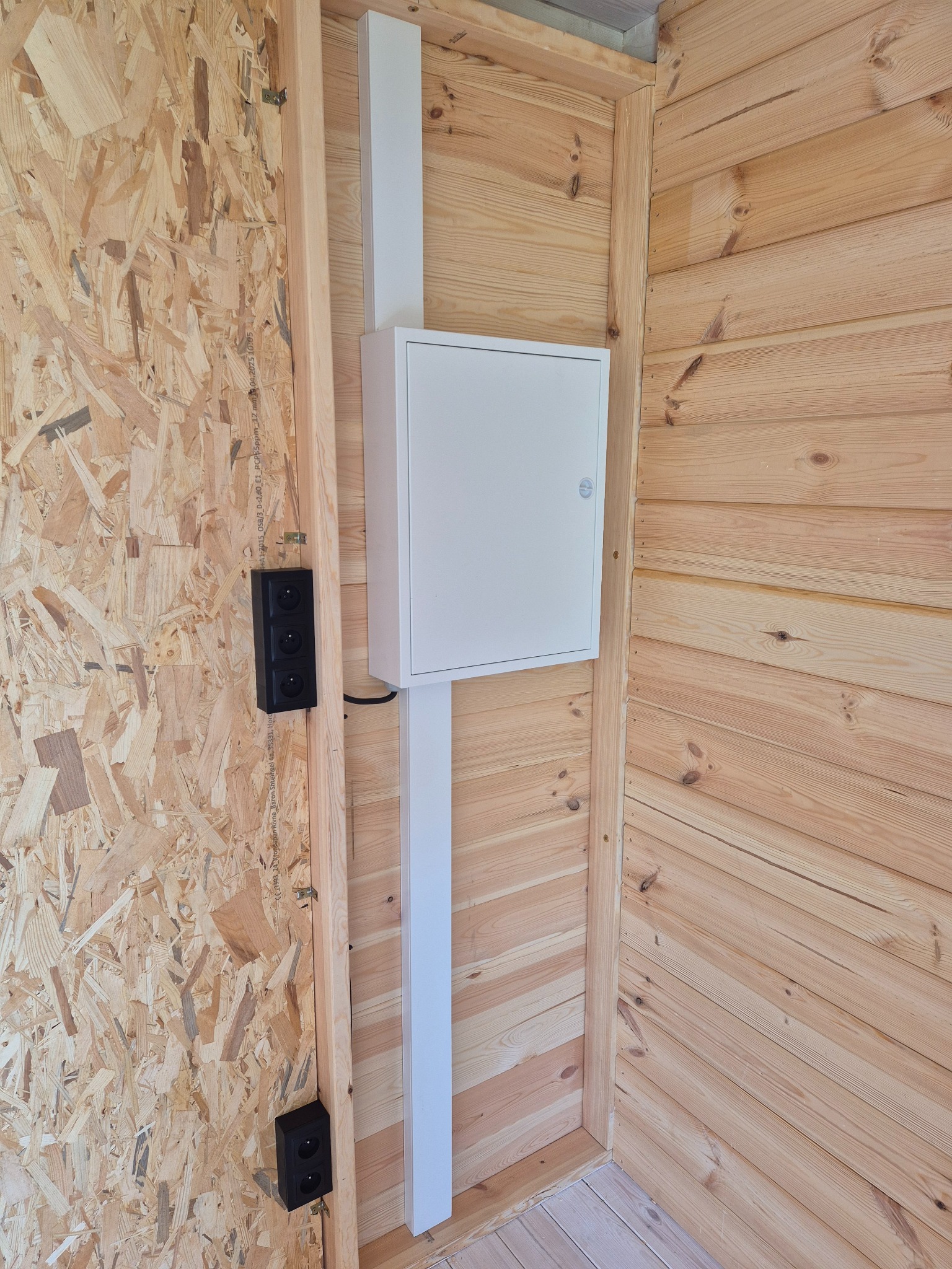 Wnętrze drewnianego domu z zamontowaną białą skrzynką elektryczną i czarnymi gniazdkami. Widoczne płyty OSB i boazeria. Ujęcie z perspektywy narożnika.
