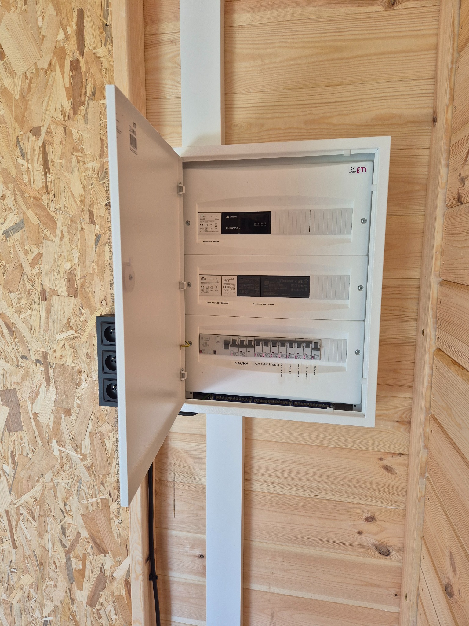 Otwarta, biała skrzynka rozdzielcza ETI z bezpiecznikami na drewnianej ścianie, obok czarne gniazdka elektryczne. Widoczne napisy 'SAUNA' i 'GN 1 GN 2 GN 3'.