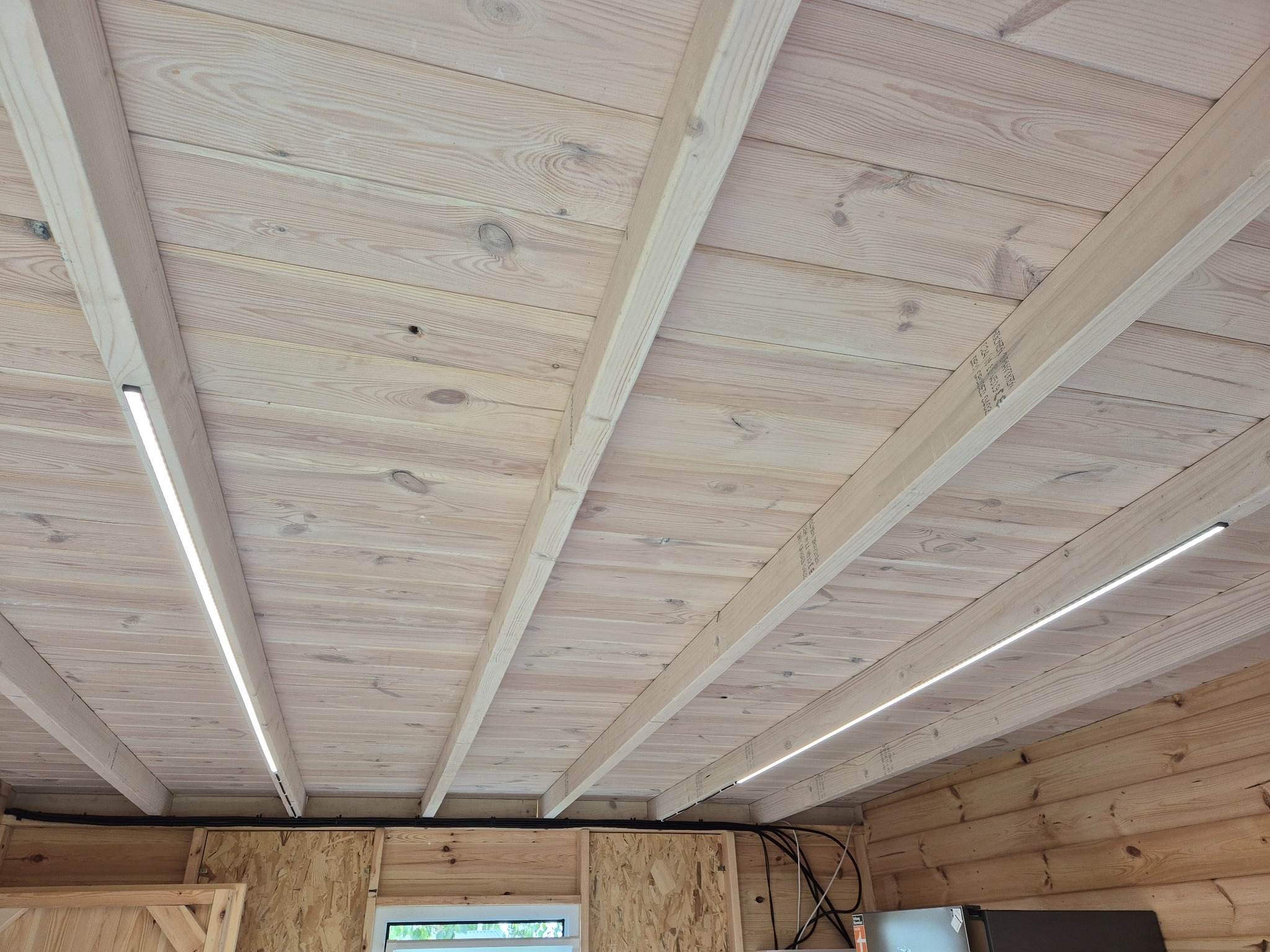 Drewniany sufit z zamontowanym oświetleniem LED wzdłuż belek. Widoczne kable i fragment ściany z płyty OSB. Ujęcie z dołu, szeroki kąt.