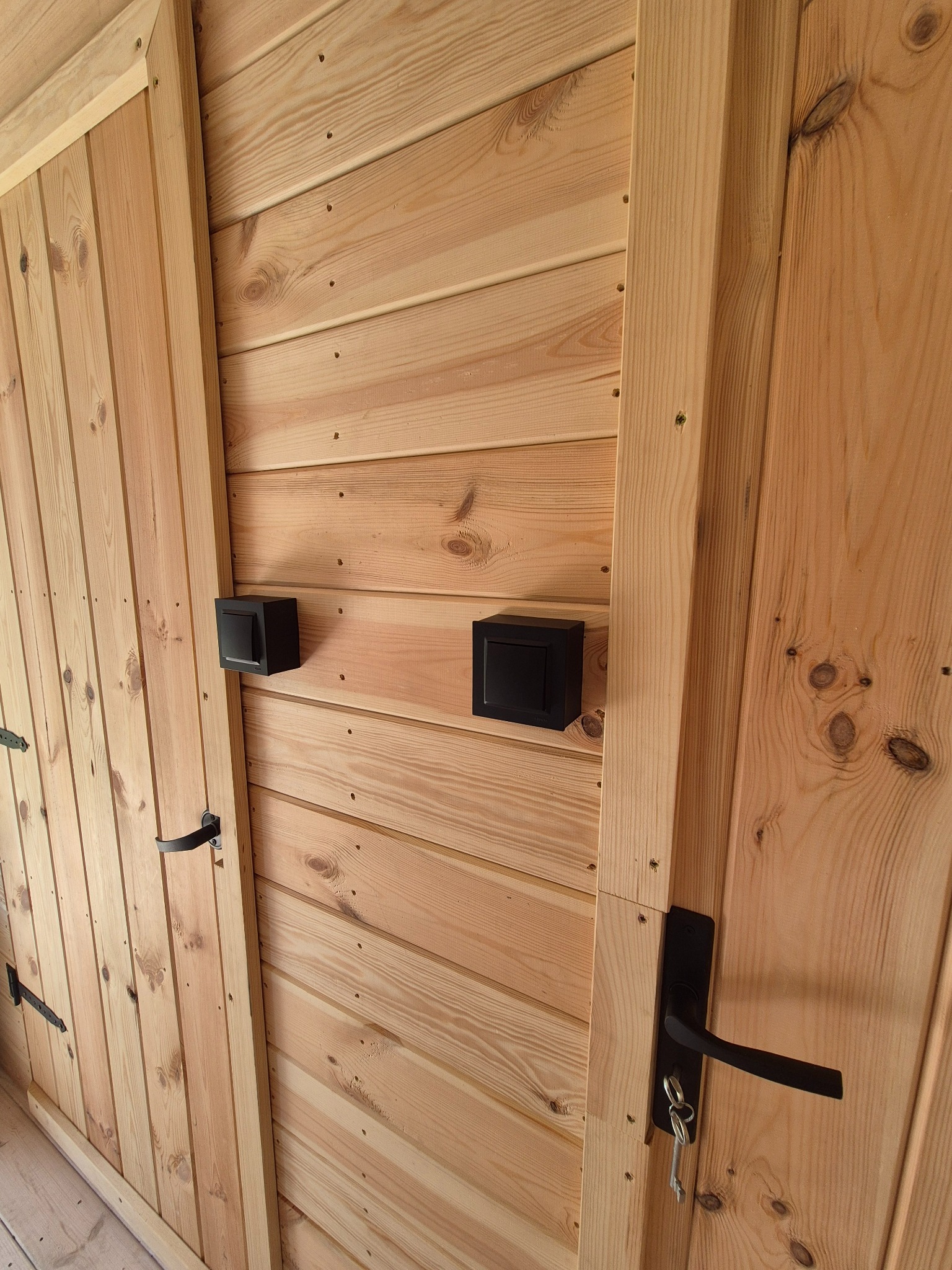 Dwa czarne, kwadratowe czujniki alarmowe zamontowane na drewnianej ścianie obok drzwi z czarną klamką i kluczami. Widoczne drewniane deski i faktura drewna.