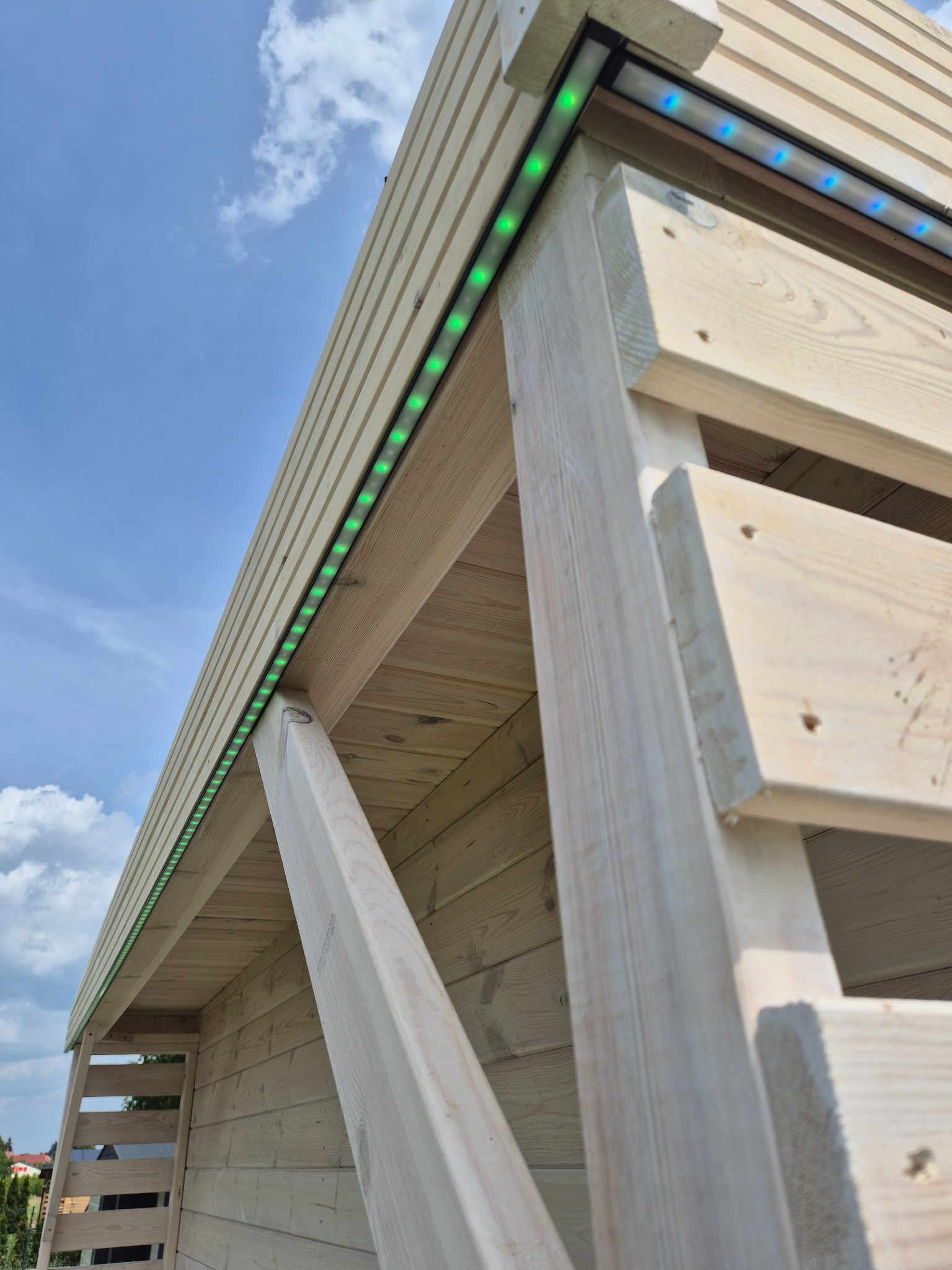 Drewniana konstrukcja z oświetleniem LED w kolorze zielonym i niebieskim, zamontowanym pod dachem, na tle błękitnego nieba z chmurami. Ujęcie z dołu.