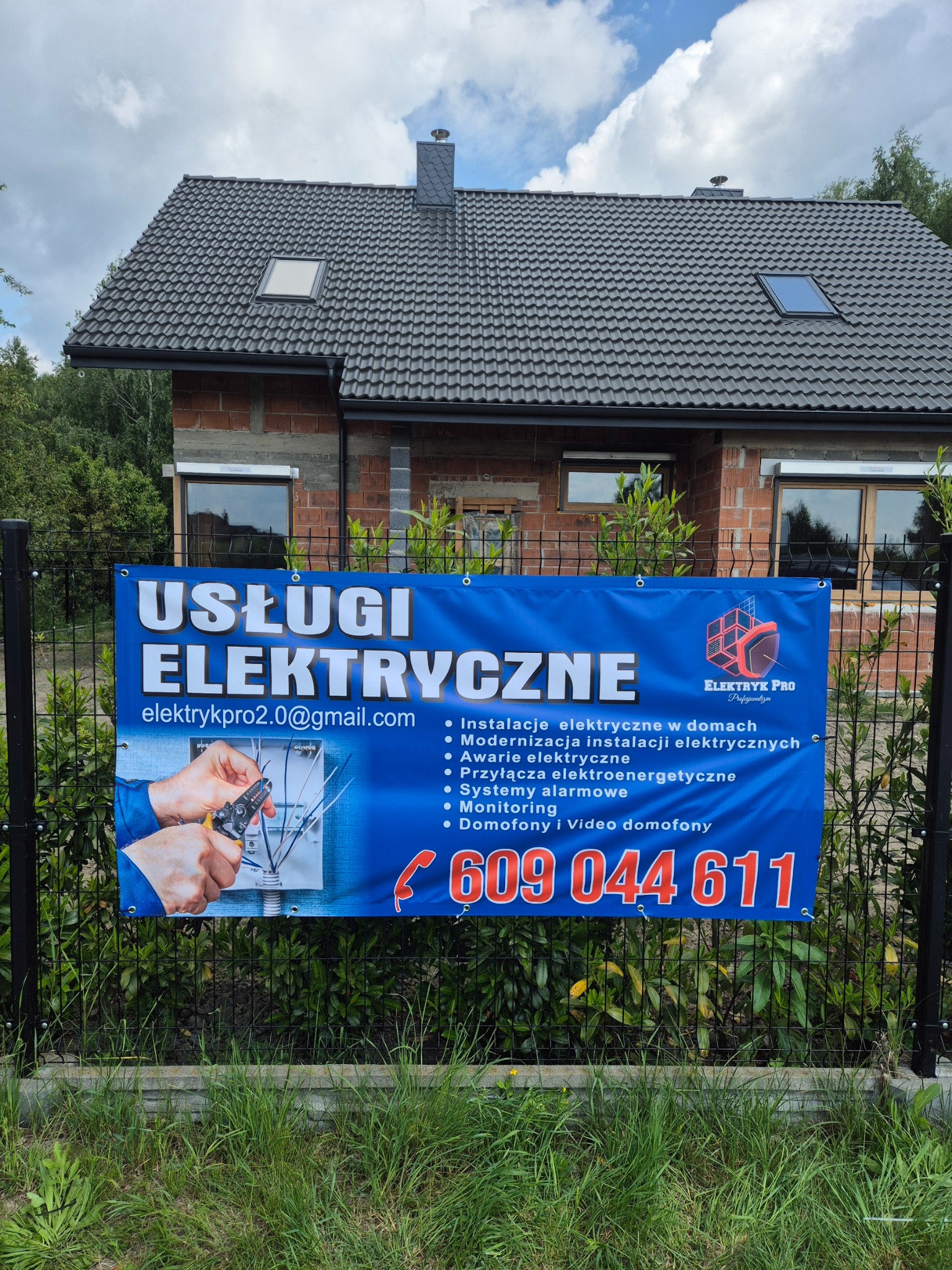 Baner reklamowy usług elektrycznych na tle domu w budowie. Oferta obejmuje instalacje, modernizacje, awarie, przyłącza, alarmy, monitoring i domofony. Kontakt: elektrykpro2.0@gmail.com.