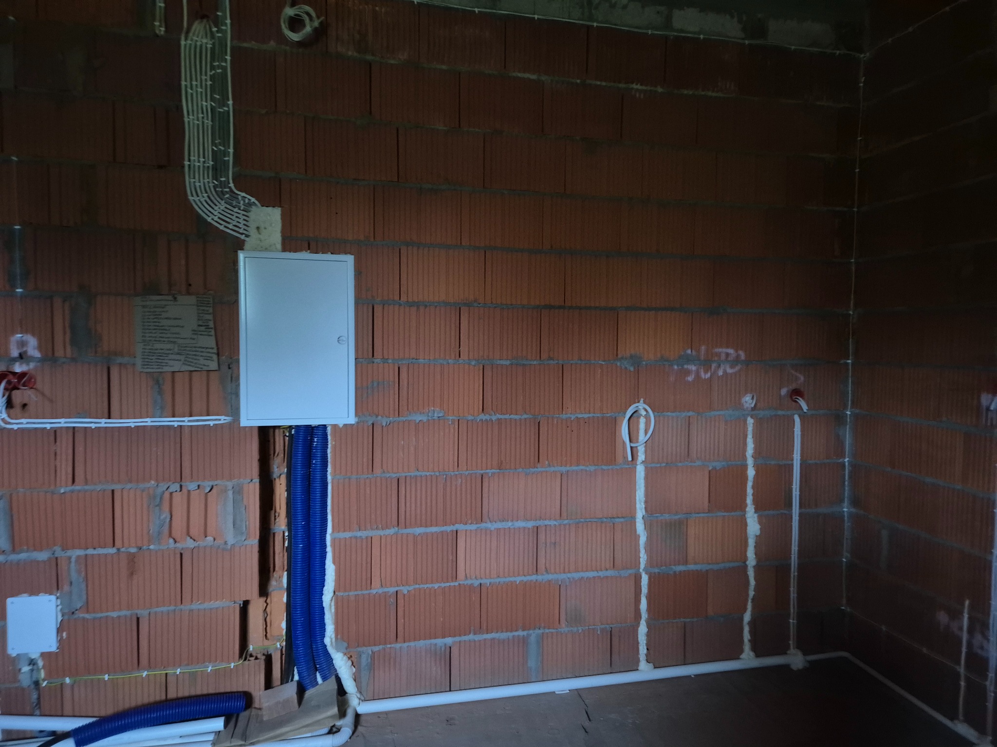 Rozdzielnia elektryczna z niebieskimi peszlami na ścianie z czerwonej cegły. Widoczne kable i przewody elektryczne. Instalacja w trakcie realizacji.