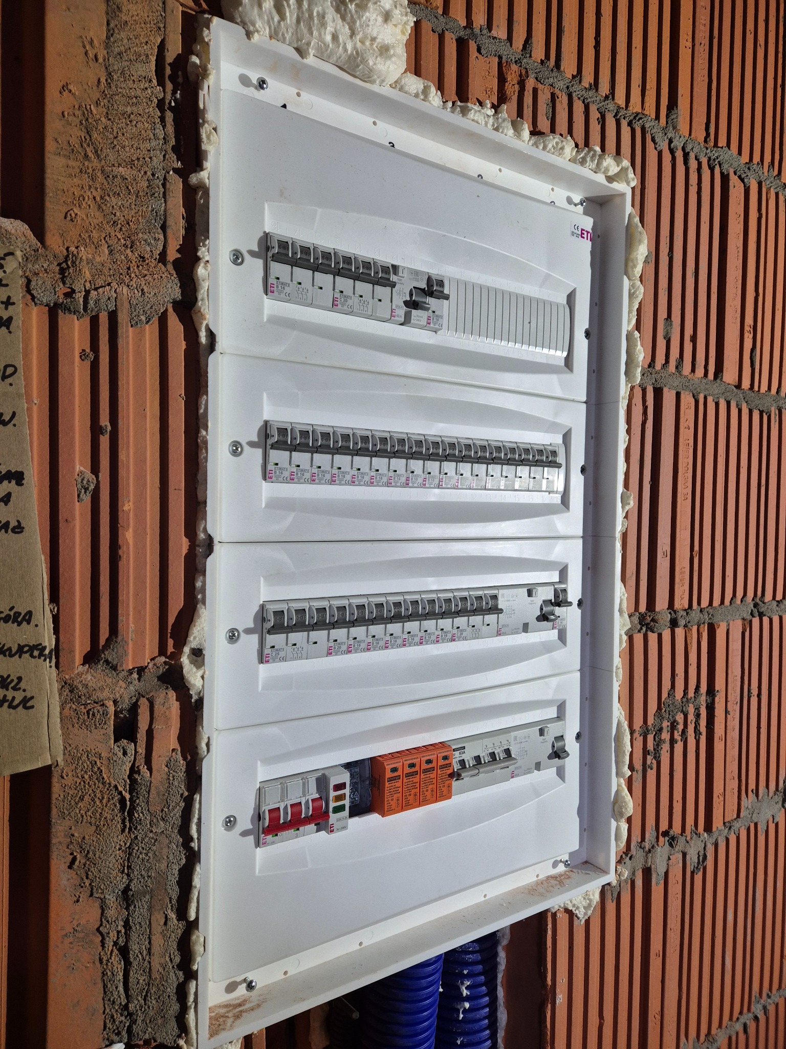 Rozdzielnica elektryczna z zabezpieczeniami, zamontowana na ceglanej ścianie z pianką montażową. Widoczne bezpieczniki i przewody. Instalacja w trakcie prac wykończeniowych.