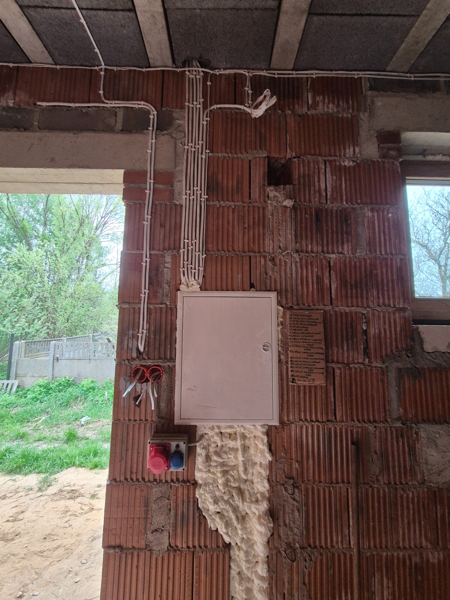 Rozprowadzenie przewodów elektrycznych w peszlach na ceglanej ścianie w surowym stanie, z zamontowaną skrzynką rozdzielczą i gniazdem siłowym. Widoczne kable i pianka montażowa.