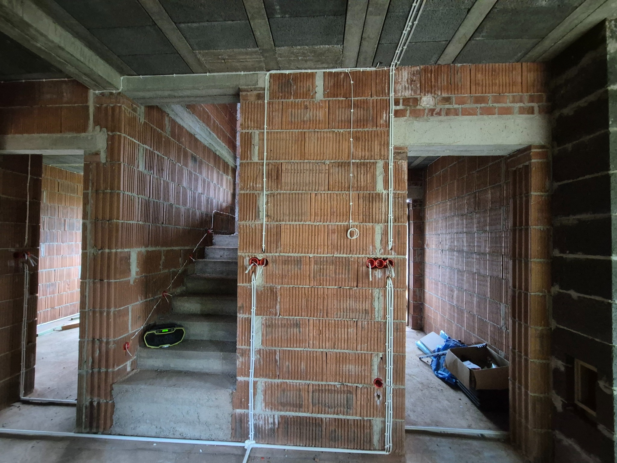 Instalacja elektryczna w surowym budynku z cegły: białe kable prowadzone po ścianach, widoczne puszki elektryczne, surowy betonowy strop, schody w tle.