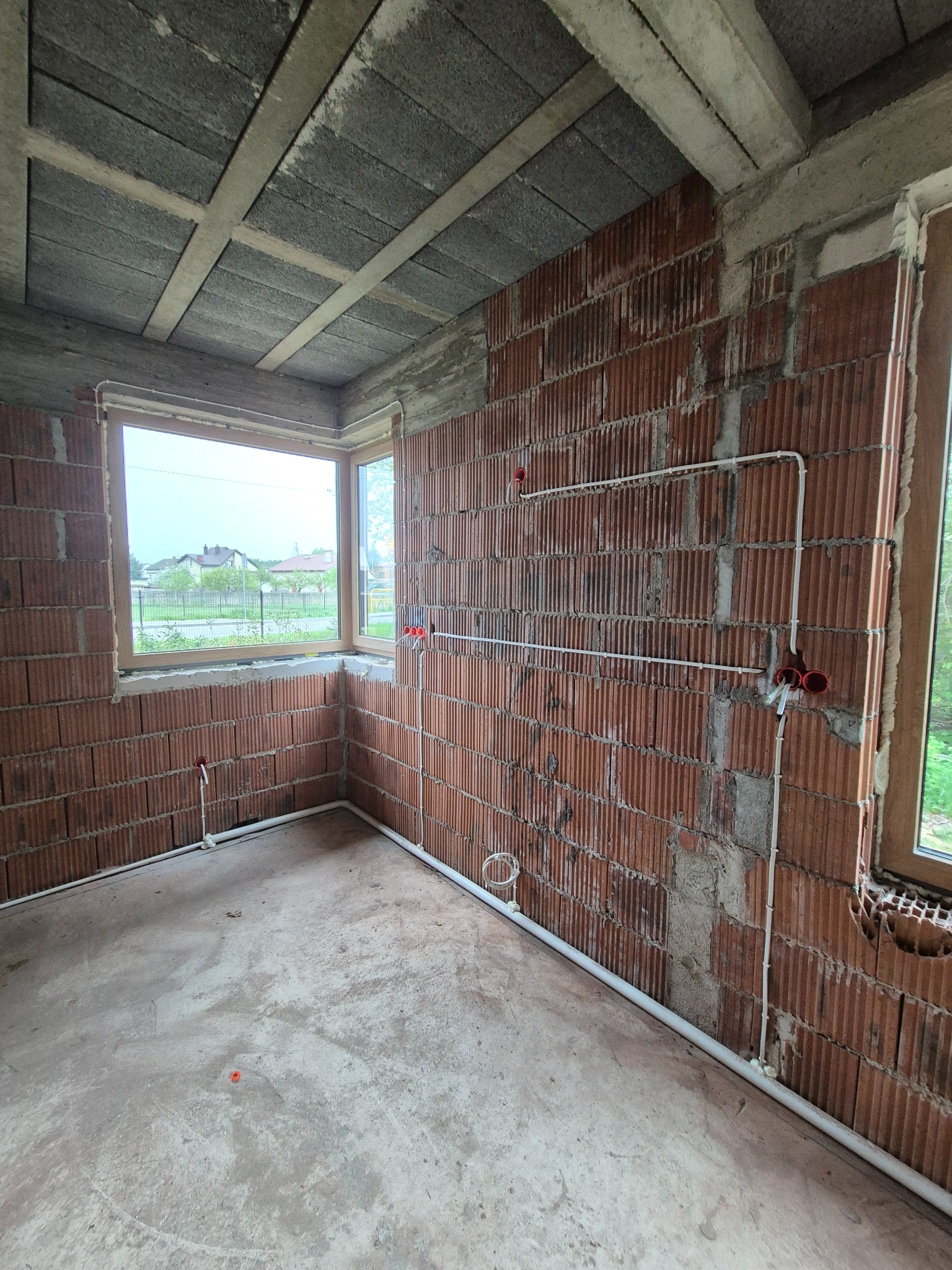 Wnętrze w trakcie prac: ściany z cegły dziurawki, białe rury instalacji elektrycznej prowadzone po ścianach, widoczne puszki elektryczne, surowy beton na podłodze.