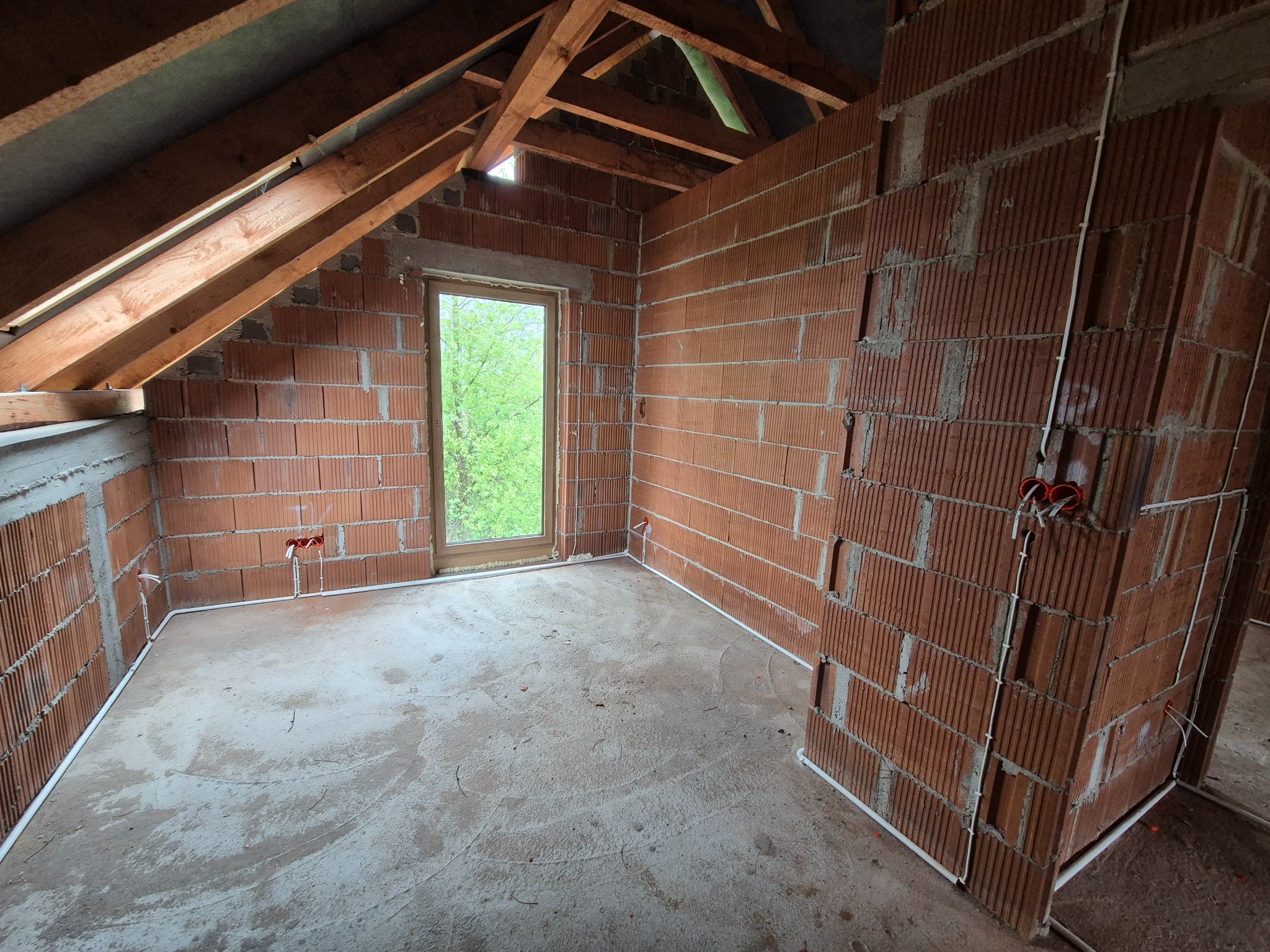 Surowy stan poddasza z ceglanymi ścianami i widoczną instalacją elektryczną w peszlach. Okno z widokiem na zieleń. Drewniana konstrukcja dachu.