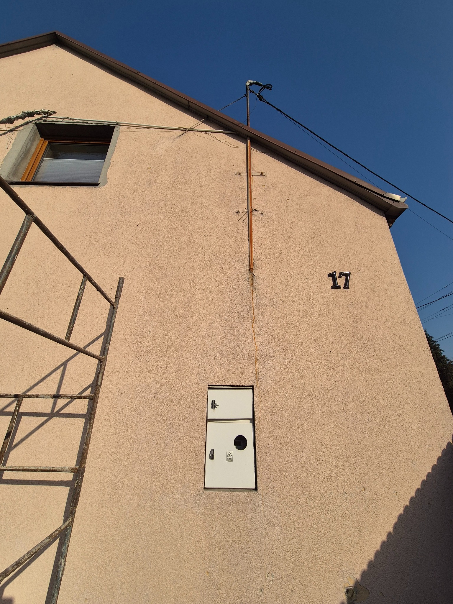 Elewacja domu z numerem 17, skrzynką elektryczną i rurą pionową. Widoczne przewody, okno i fragment rusztowania. Ujęcie z dołu, błękitne niebo w tle.