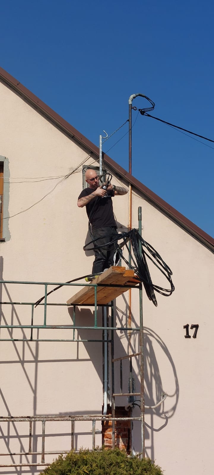Elektryk na rusztowaniu montuje kable na elewacji budynku z numerem 17. Widoczne narzędzia i instalacja antenowa. Słoneczny dzień, błękitne niebo.